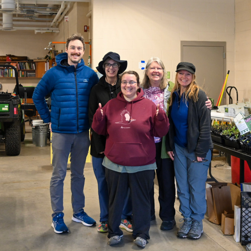 Five volunteers standing together in an indoor workspace with tables of native plants and supplies behind them.