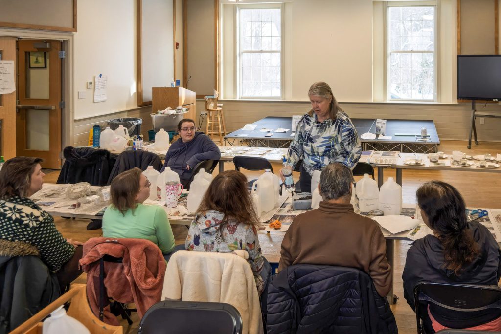 Presenter demonstrates seed sowing techniques to seated workshop participants gathered around tables with milk jugs and supplies