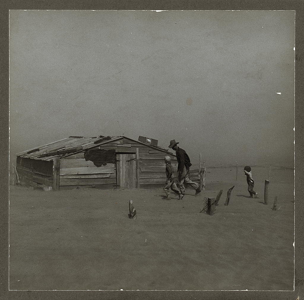 Black-and-white photograph of a farmer and two children walking into a dust storm in Cimarron County, Oklahoma. The image documents the severe soil erosion and wind-driven dust conditions of the Dust Bowl era.